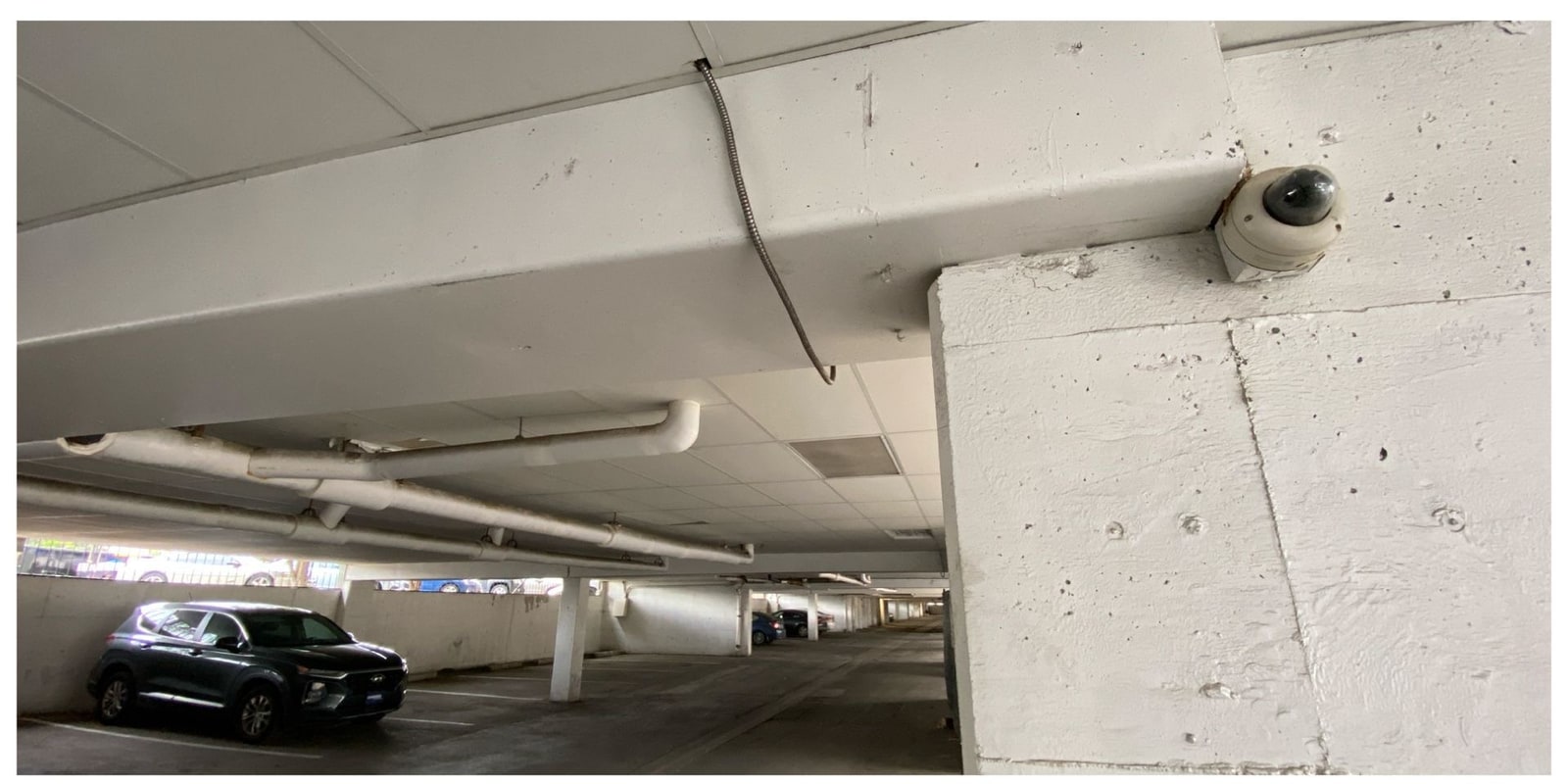 Dome camera mounted on a concrete beam above a covered parking aisle with a parked SUV