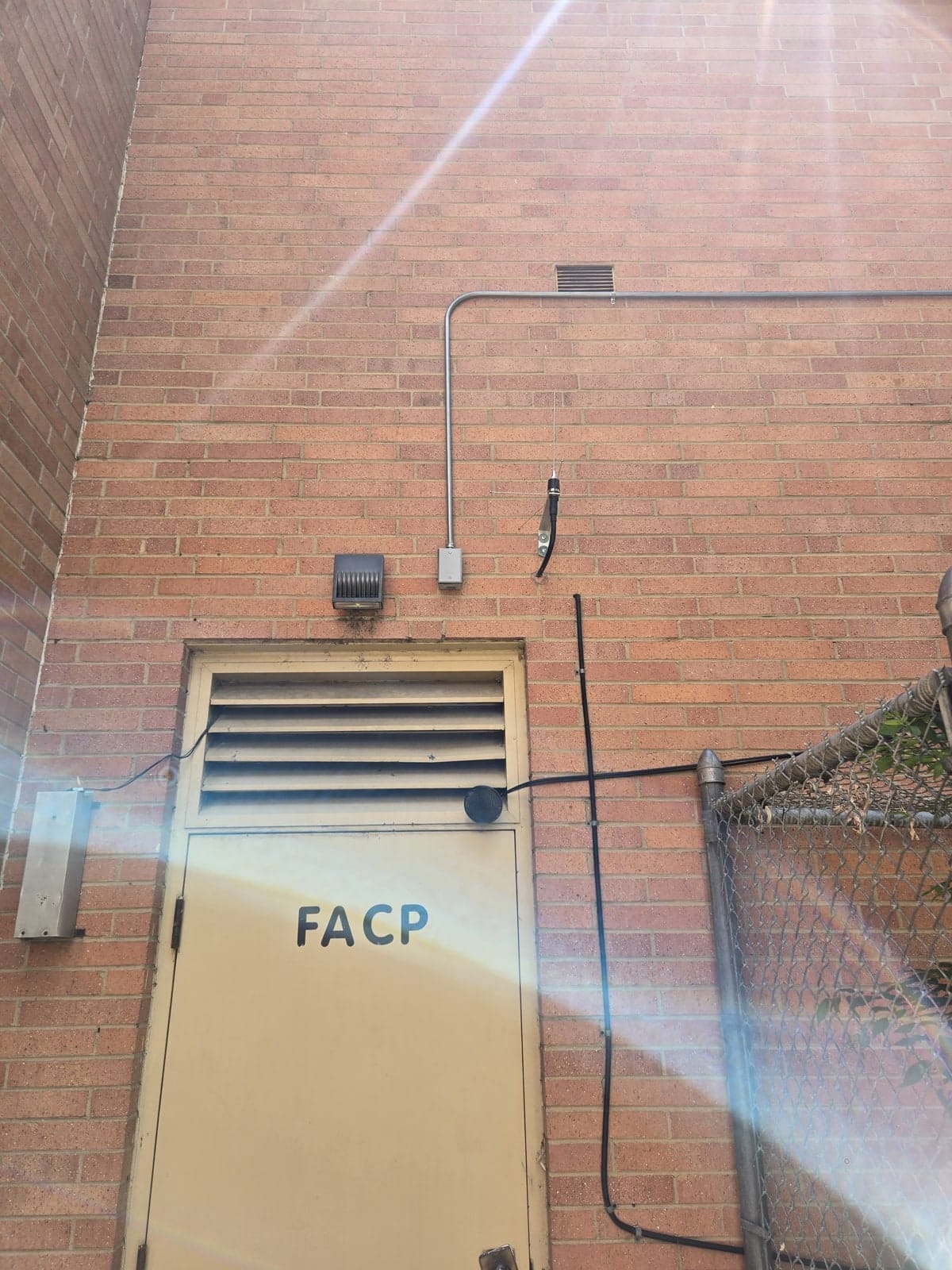 Brick wall with a fire alarm control panel door, surface conduit and chain-link fence